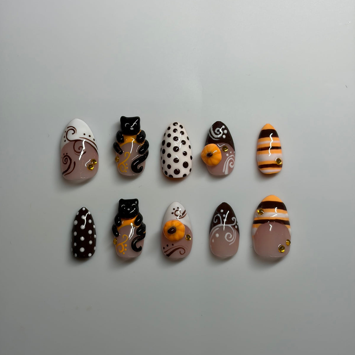 A set of 10 press-on nails arranged in two rows of five on a plain light gray background. The nails feature various Halloween-themed designs including black cats with gold accents, white polka dots, orange pumpkins, and striped patterns.
