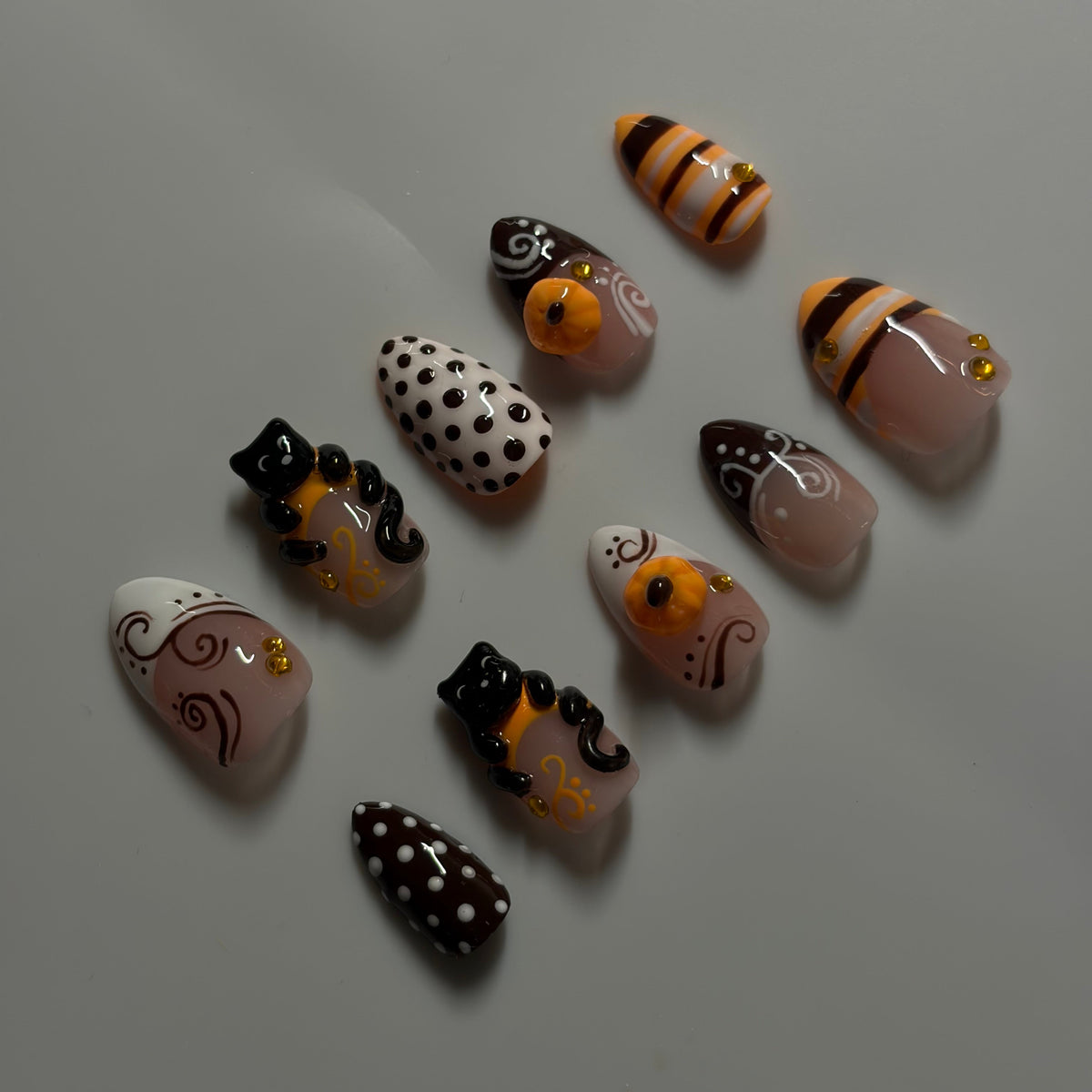 A set of artificial press-on nails arranged on a plain light gray surface. The nails feature Halloween-themed designs including black cats with gold accents, white and black polka dots, orange and black stripes, and pale pink nails with gold swirl details.